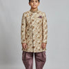 Boy wearing cream and gold floral sherwani with purple dhoti pants and traditional juttis on gray background