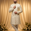 Man wearing a cream sherwani with intricate patterns against a beige curtain backdrop.