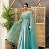 Woman wearing a teal embroidered crop top and flowing skirt with sheer sleeves, standing indoors by a decorative wooden screen and plant