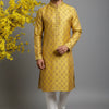 Man wearing golden yellow traditional Indian kurta with white churidar and jutti, standing by yellow flowers