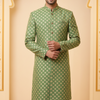 Man wearing a green patterned kurta with white pants against a beige wall.