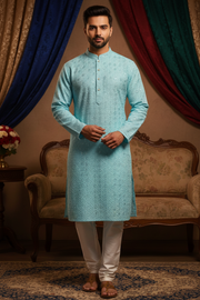 A man modeling a light blue embroidered kurta with white trousers, standing in a traditional indoor setting with draped blue, red, and green curtains and a vintage sofa.