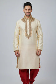 Man wearing cream-colored embroidered Indian kurta with red churidar pants, standing against gray background