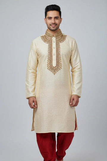 Man wearing cream-colored embroidered Indian kurta with red churidar pants, standing against gray background