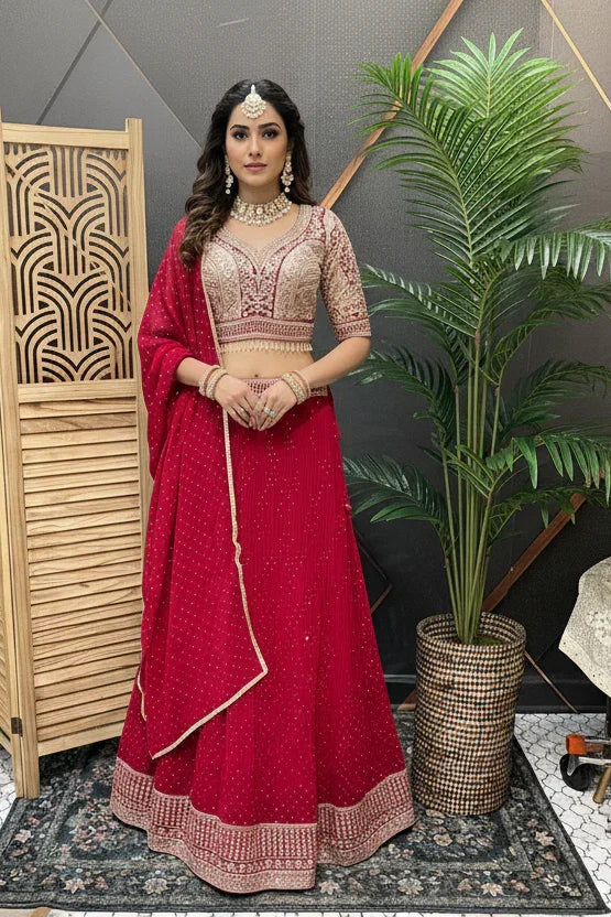 Woman in red and beige embroidered Indian lehenga with jewelry, standing indoors by plant and wooden screen