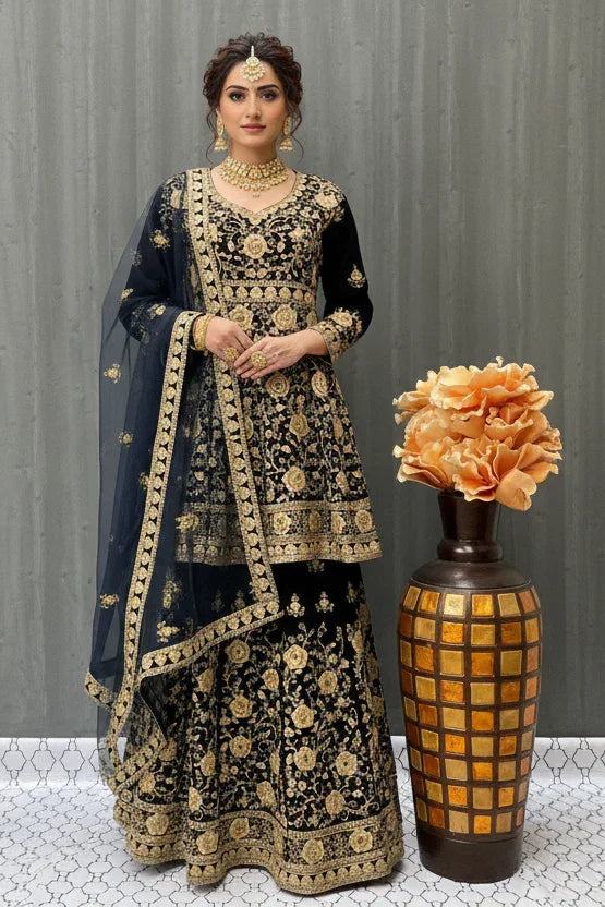 Woman in black and gold embroidered Indian ethnic lehenga outfit with matching jewelry, standing beside decorative vase with orange flowers