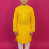 Young boy in bright yellow Indian ethnic kurta with white churidar, standing barefoot against pink background