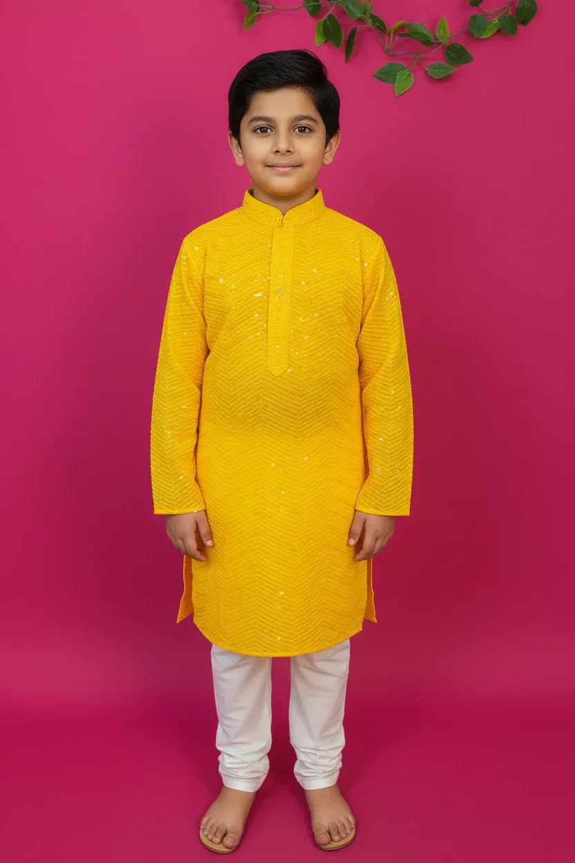 Young boy in bright yellow Indian ethnic kurta with white churidar, standing barefoot against pink background