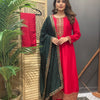 Woman in red Indian ethnic suit with intricate embroidery, black embellished dupatta, standing indoors by wooden screen and plant