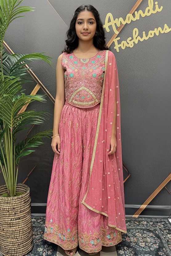 Woman in a pink traditional outfit standing next to a plant with 'Anandi Fashions' branding in the background.