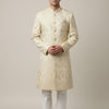 A man modeling a traditional Indian sherwani long coat in cream beige with gold floral brocade, paired with white trousers and beige shoes, against a solid light beige background.