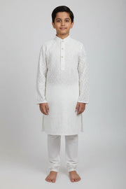 Boy wearing white embroidered Indian kurta with churidar pants, standing barefoot on plain background