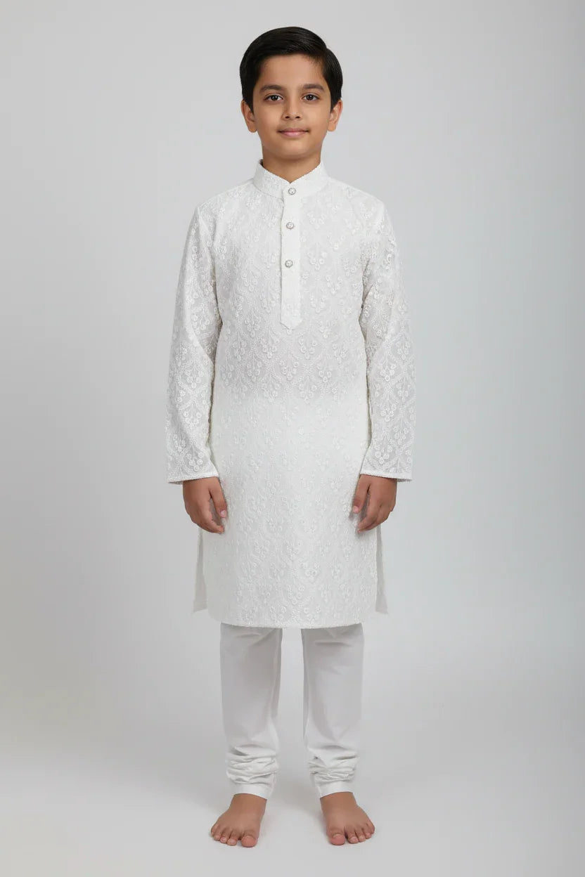 Boy wearing white embroidered Indian kurta with churidar pants, standing barefoot on plain background