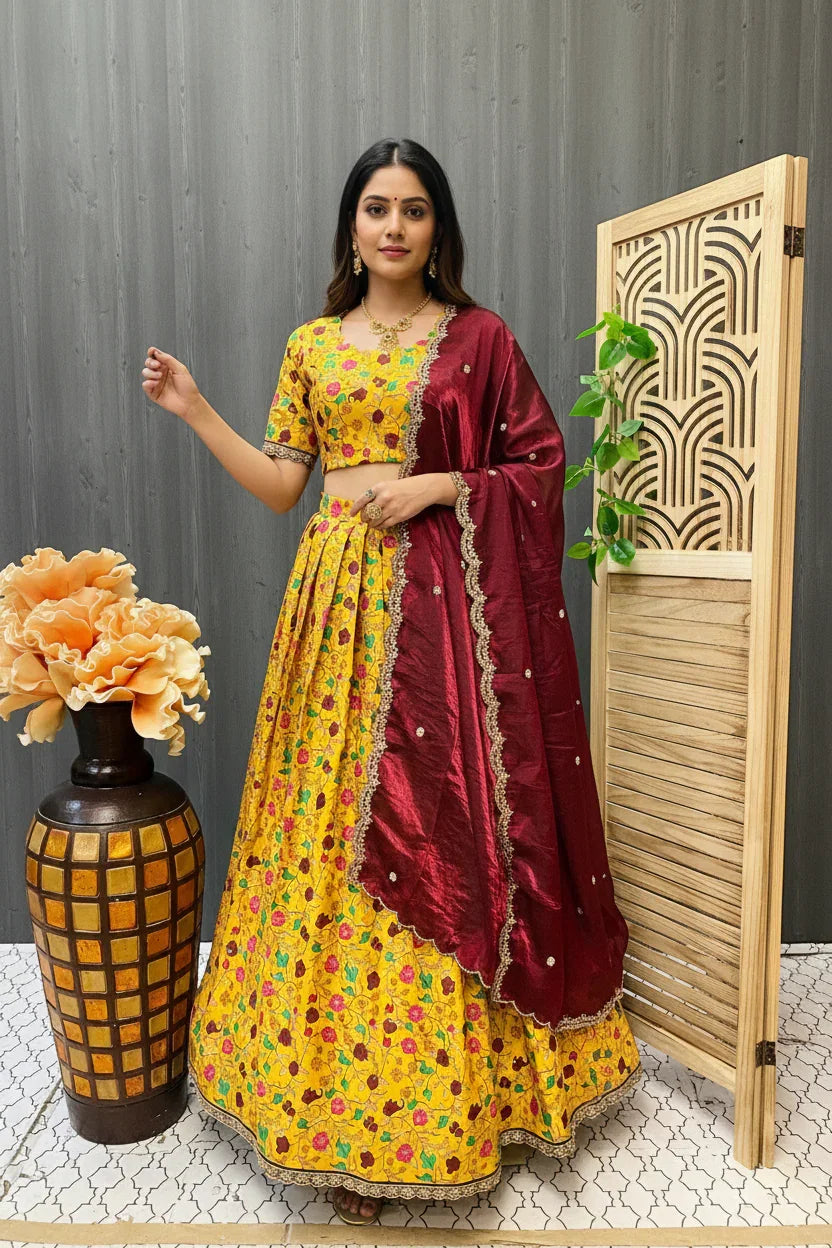 Woman wearing yellow floral lehenga with maroon dupatta, standing indoors near decorative vase and wooden screen