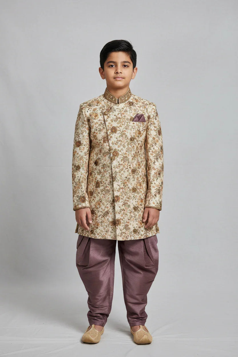 Boy wearing cream and gold floral sherwani with purple dhoti pants and traditional juttis on gray background