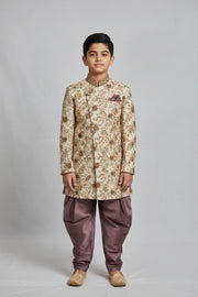 Boy wearing cream and gold floral sherwani with purple dhoti pants and traditional juttis on gray background