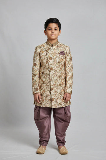 Boy wearing cream and gold floral sherwani with purple dhoti pants and traditional juttis on gray background