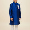 Young boy wearing a royal blue silk kurta and white churidar pants, standing barefoot on beige background