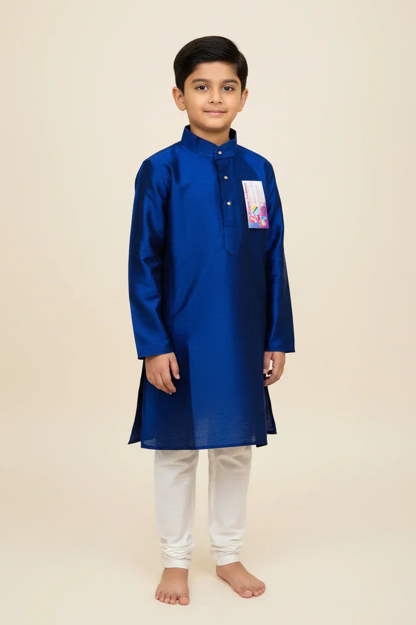 Young boy wearing a royal blue silk kurta and white churidar pants, standing barefoot on beige background