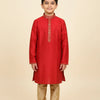 Young boy wearing red Indian kurta with golden embroidery and beige churidar pants, standing barefoot