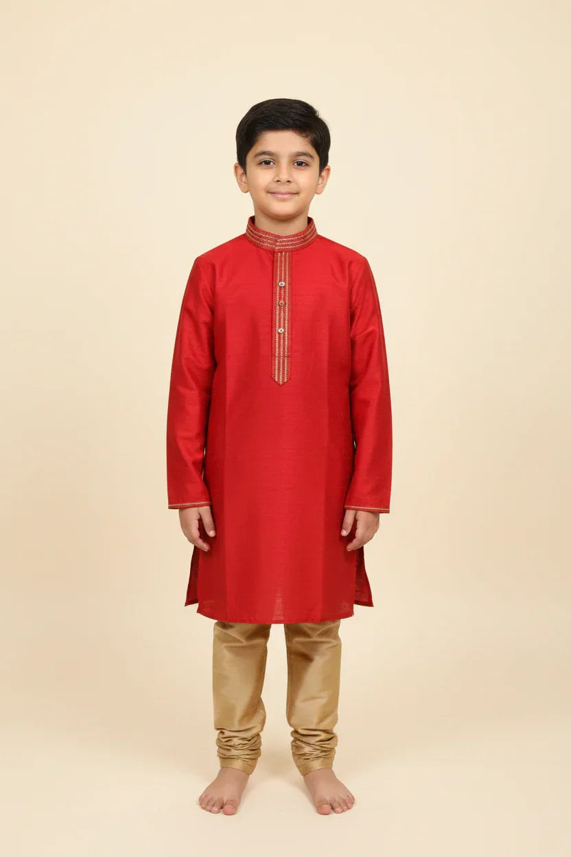 Young boy wearing red Indian kurta with golden embroidery and beige churidar pants, standing barefoot