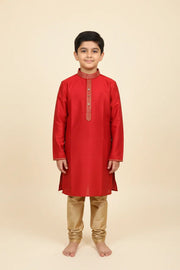 Young boy wearing red Indian kurta with golden embroidery and beige churidar pants, standing barefoot