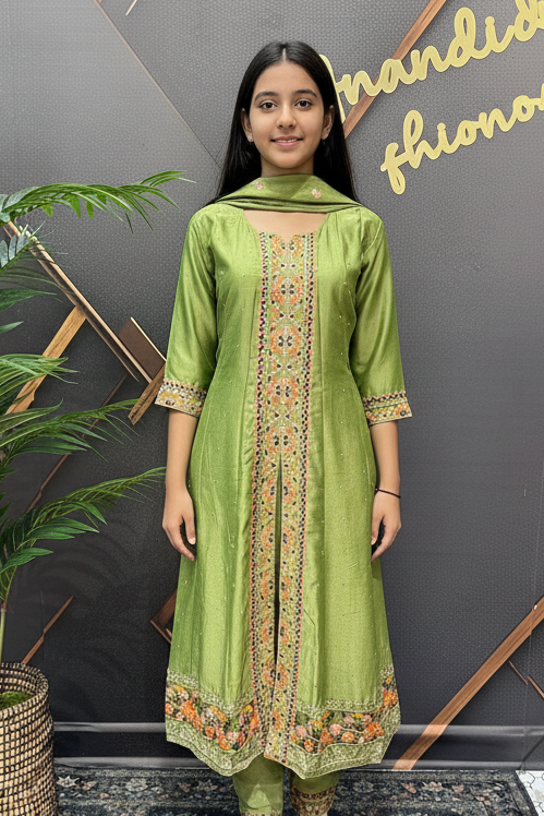 Woman in a green traditional outfit standing next to a potted plant against a decorative wall.