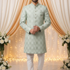 Man wearing a light green sherwani with white pants against a decorated backdrop with lights and flowers.
