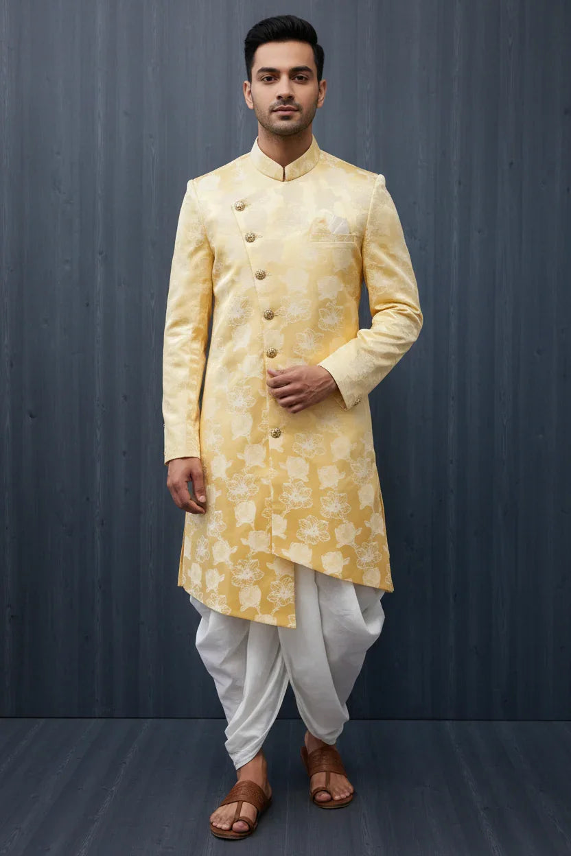 Man wearing a yellow floral brocade sherwani with white dhoti pants and brown sandals in front of dark background