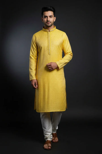 Man wearing a yellow embroidered Indian kurta with white churidar pants and brown sandals on dark background
