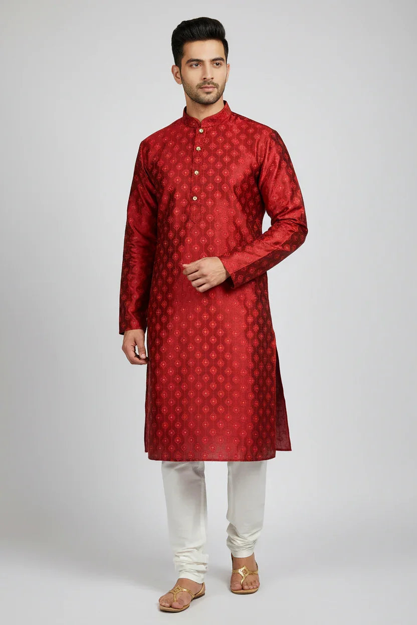 Man wearing red patterned silk kurta with white churidar and traditional sandals against grey background