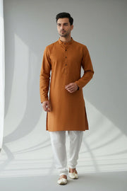 Man wearing rust orange Indian kurta with white churidar pants and traditional shoes in minimalist setting
