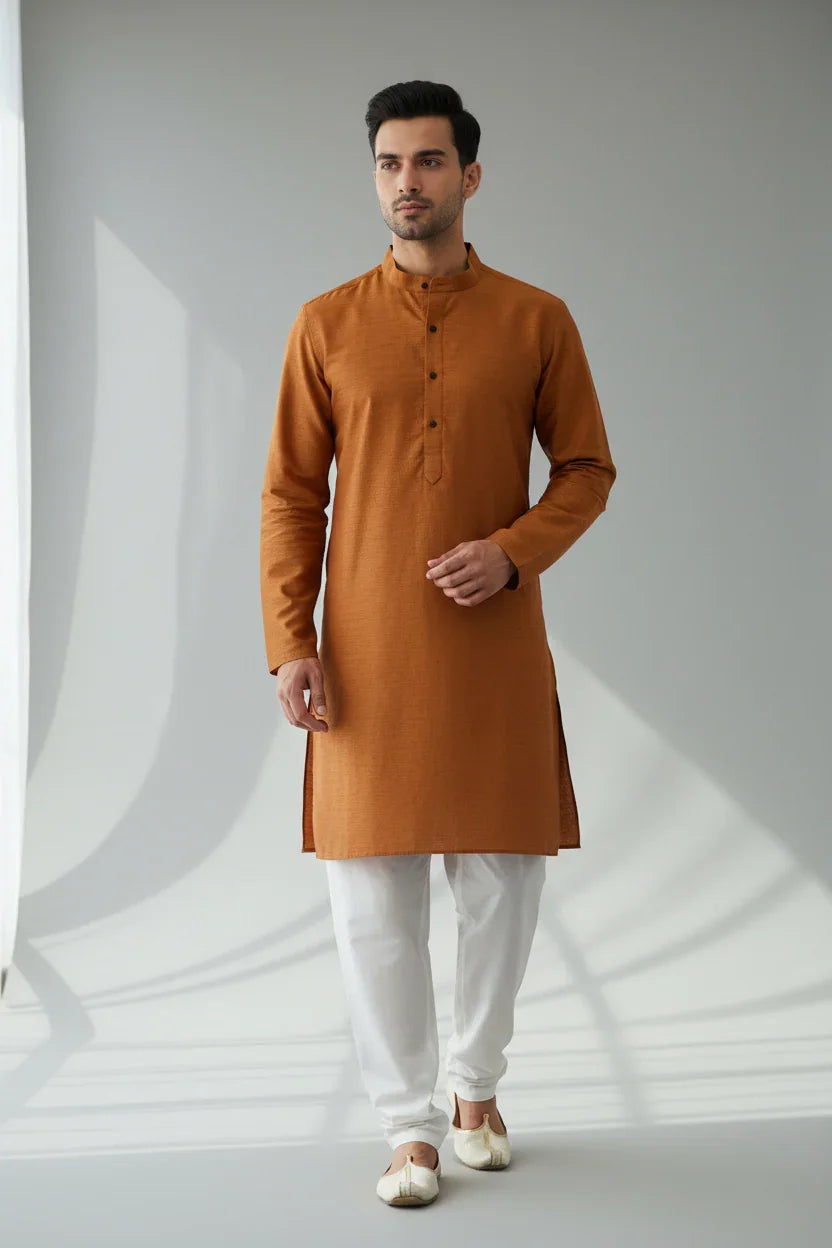 Man wearing rust orange Indian kurta with white churidar pants and traditional shoes in minimalist setting