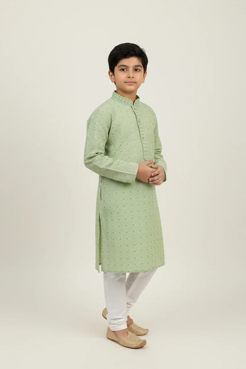 Boy in light green embroidered Indian kurta with white churidar and traditional shoes, studio background