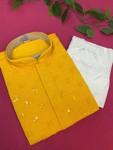 Folded yellow embroidered Indian ethnic kurta with white churidar pants on pink fabric background, decorative green leaves