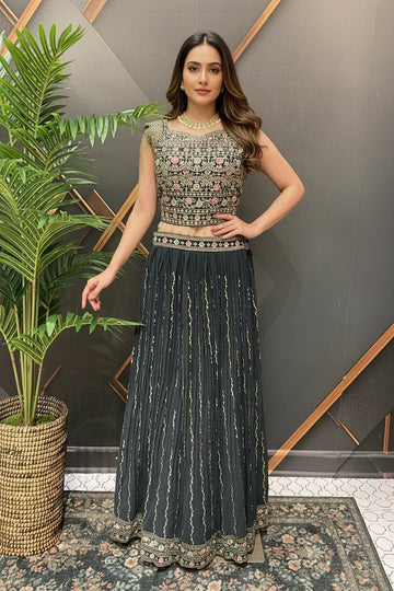 Woman wearing black embroidered ethnic lehenga choli with floral patterns, standing indoors near potted plant