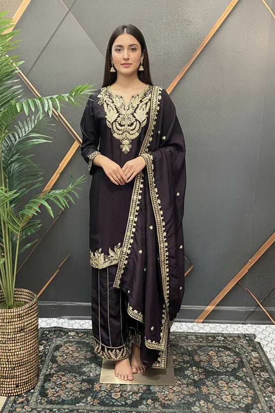 Woman in black Indian ethnic wear with gold embroidery, standing barefoot on patterned rug near plant