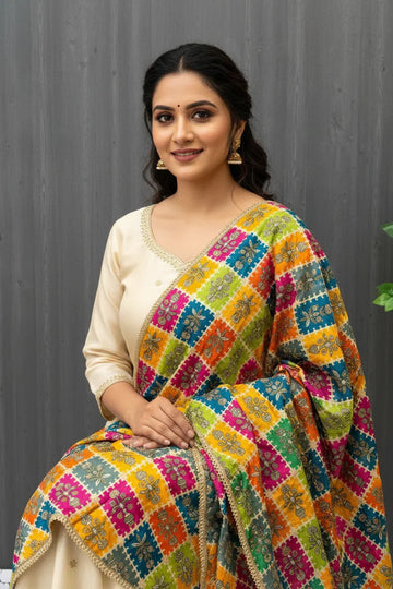 Woman in cream ethnic wear with vibrant multicolor embroidered dupatta sitting against gray backdrop, wearing gold jhumka earrings