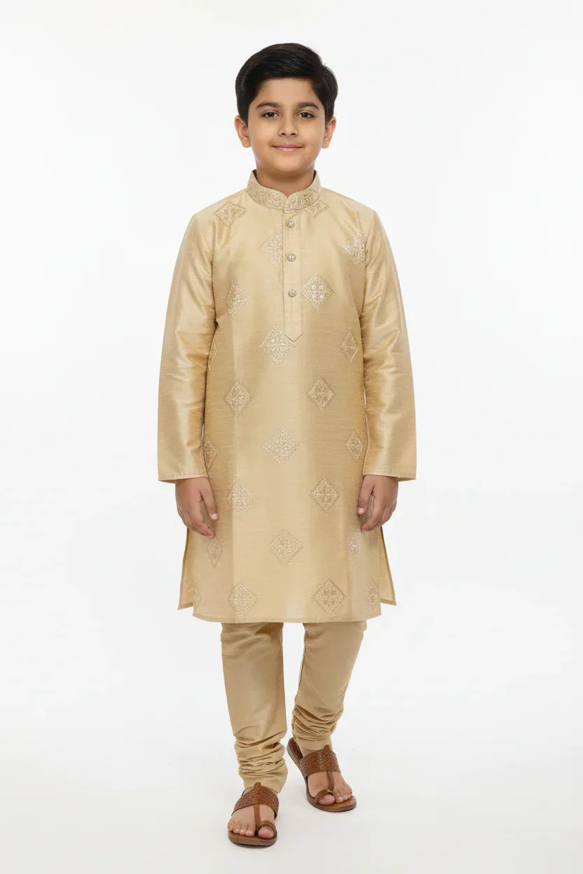Boy wearing gold embroidered kurta set with churidar and brown sandals against white background