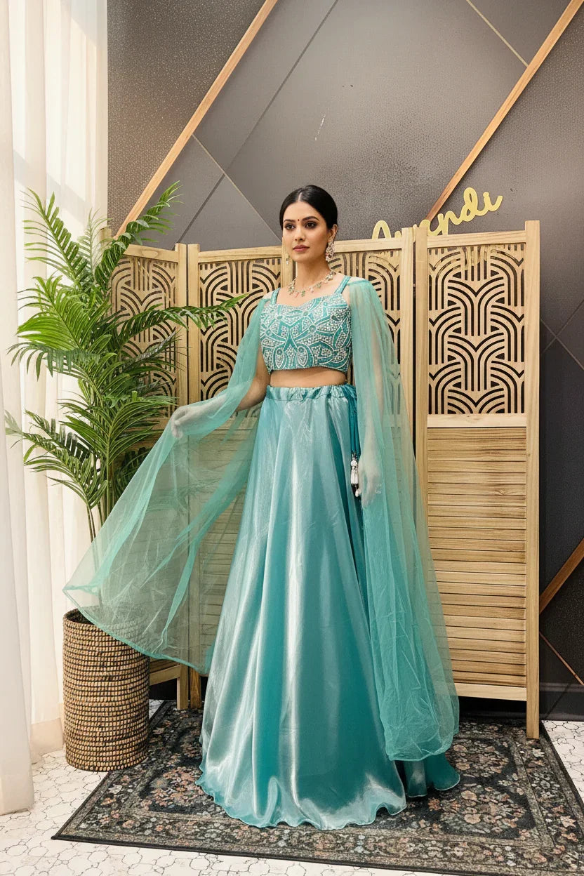 Woman wearing a teal embroidered crop top and flowing skirt with sheer sleeves, standing indoors by a decorative wooden screen and plant