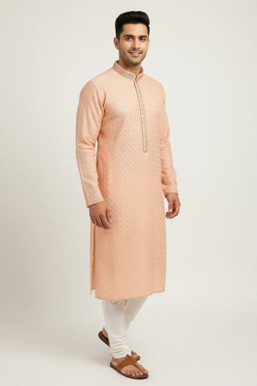 Man wearing a peach embroidered kurta with white churidar and brown sandals, standing against plain background