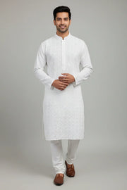 Man wearing white embroidered Indian ethnic kurta and churidar with brown traditional shoes on gray background