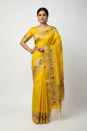 Woman wearing bright yellow traditional Indian saree with floral gold border, gold jewelry, standing against white background