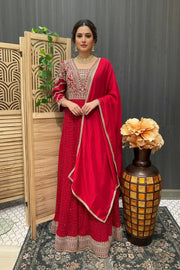 Woman in red embroidered Indian ethnic wear with matching dupatta, standing indoors near wooden screen and decorative vase