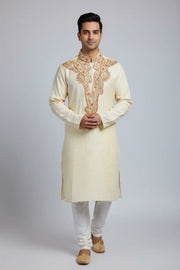 Man wearing cream embroidered Indian kurta with white churidar and traditional beige juttis on gray background