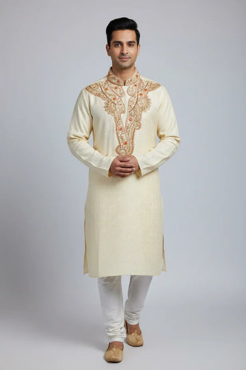 Man wearing cream embroidered Indian kurta with white churidar and traditional beige juttis on gray background