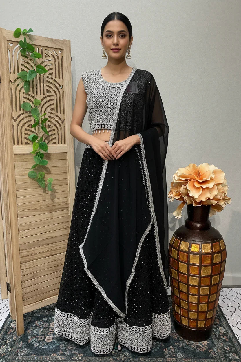 Woman in black and white embroidered Indian lehenga with sheer dupatta standing indoors near wooden screen and decorative vase