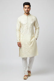 Man wearing cream-colored embroidered Indian kurta with white churidar and traditional juttis on plain background