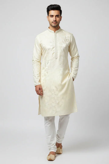 Man wearing cream-colored embroidered Indian kurta with white churidar and traditional juttis on plain background