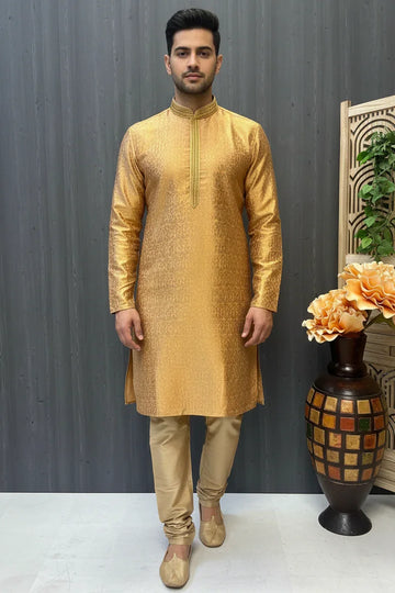 Man wearing golden Indian ethnic kurta with beige churidar and traditional shoes, standing indoors near vase and folding screen